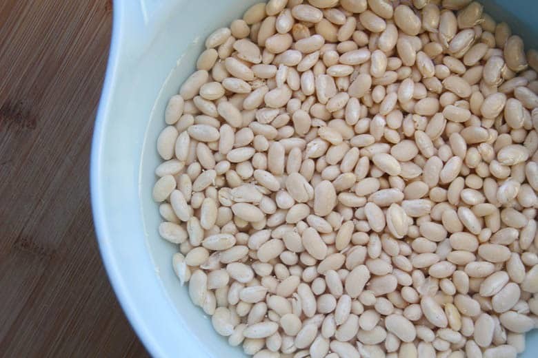 A large bowl full of dried white beans soaking in water.